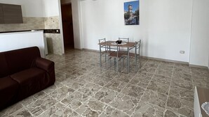 Uma sala de estar espaçosa com piso de pedra, uma mesa de jantar com cadeiras, um sofá e uma cozinha ao fundo.