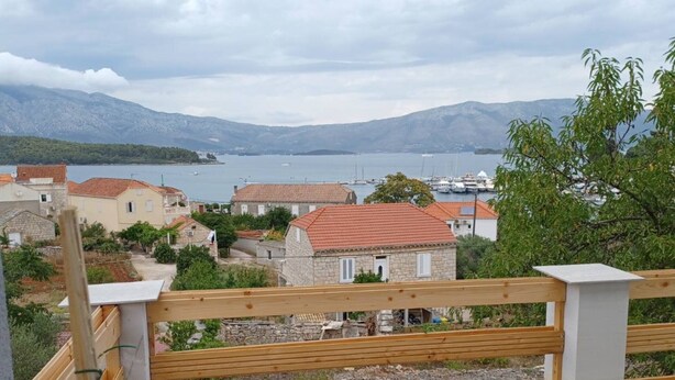 Ein Küstendorf mit Steinhäusern, einem Holzzaun und Blick auf das Meer und die Berge im Hintergrund.