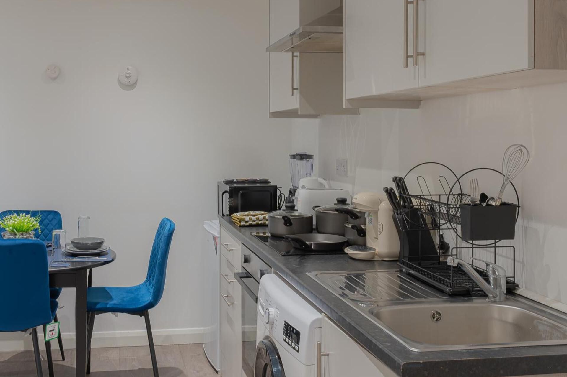 A modern kitchen with a sink, stove, and various cooking utensils. There is a blue chair and a small dining table with a vase of flowers.