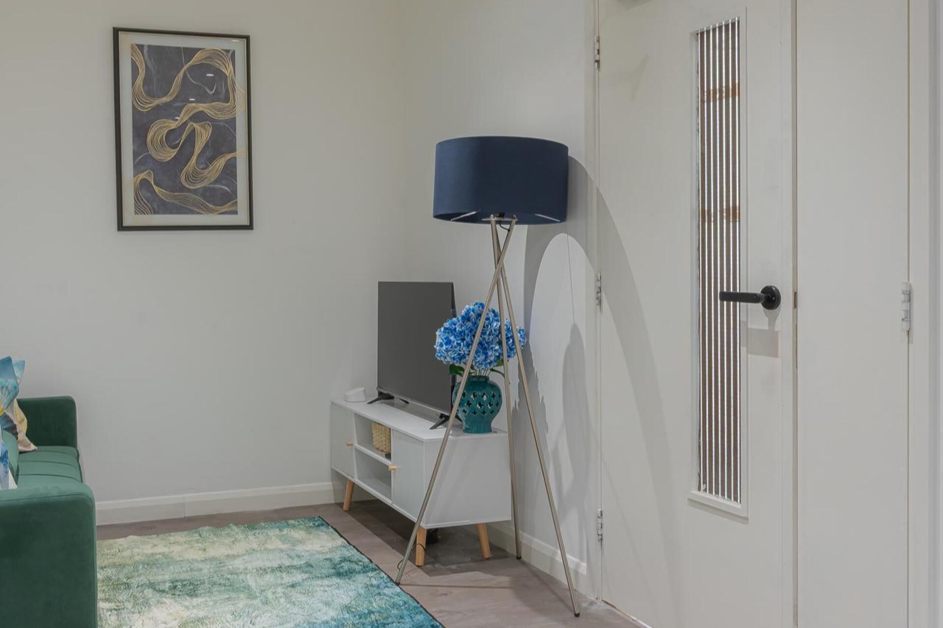 Een kamer met een witte deur, een lijst met abstract schilderij, een blauwe vloerlamp, een groene bank en een kleine tafel met een bloemenvaas.