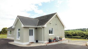 A single-story house with a grey roof and white walls, a small porch with potted plants, and a clear view of the surrounding countryside. - Farmer's Cottage (Nenagh)