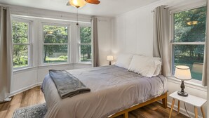 A bedroom with a bed, a ceiling fan, a nightstand with a lamp, and large windows with curtains. - Lakeside, 2-Bedroom Cottage with Fire Pit in Dickson (Dickson)