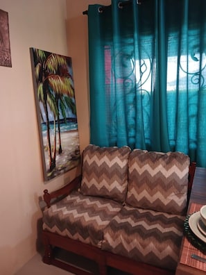 A living room with a sofa, a painting of palm trees, and teal curtains. - Casa Cicada Sleeps 4 With Balcony (Chaguanas)