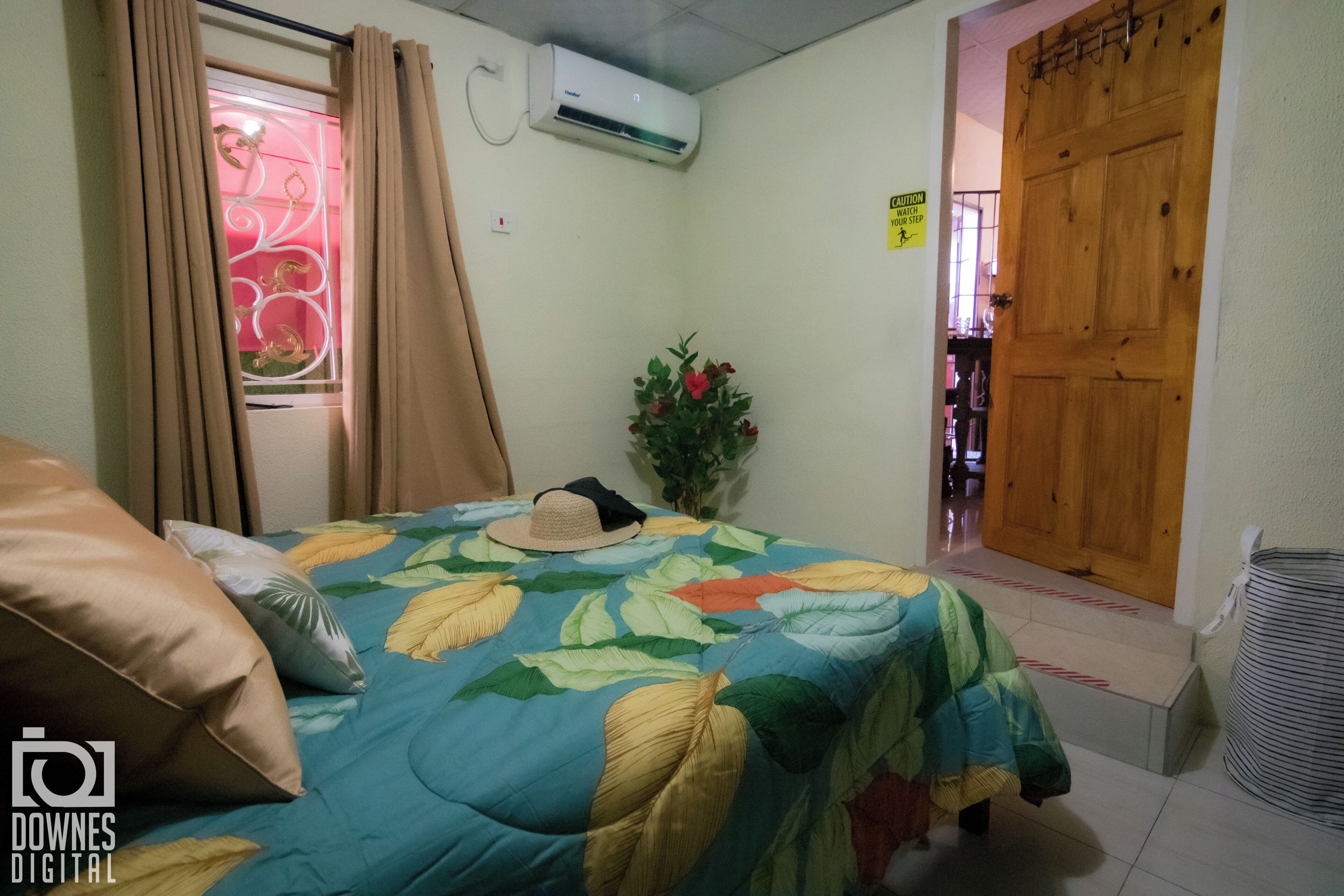 A bedroom with a bed, a window with curtains, an air conditioning unit, a wooden door, and a trash bin.