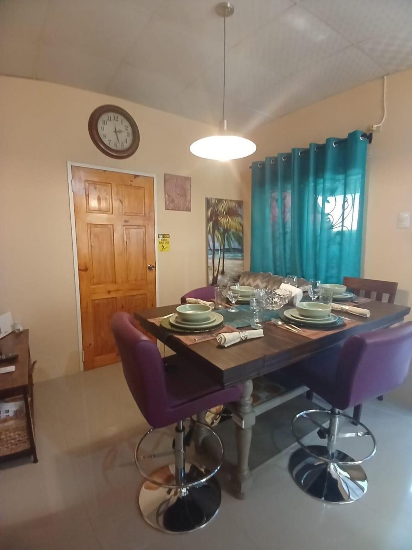 A dining area with a wooden table set for four, purple chairs, a clock on the wall, and a painting above the door.