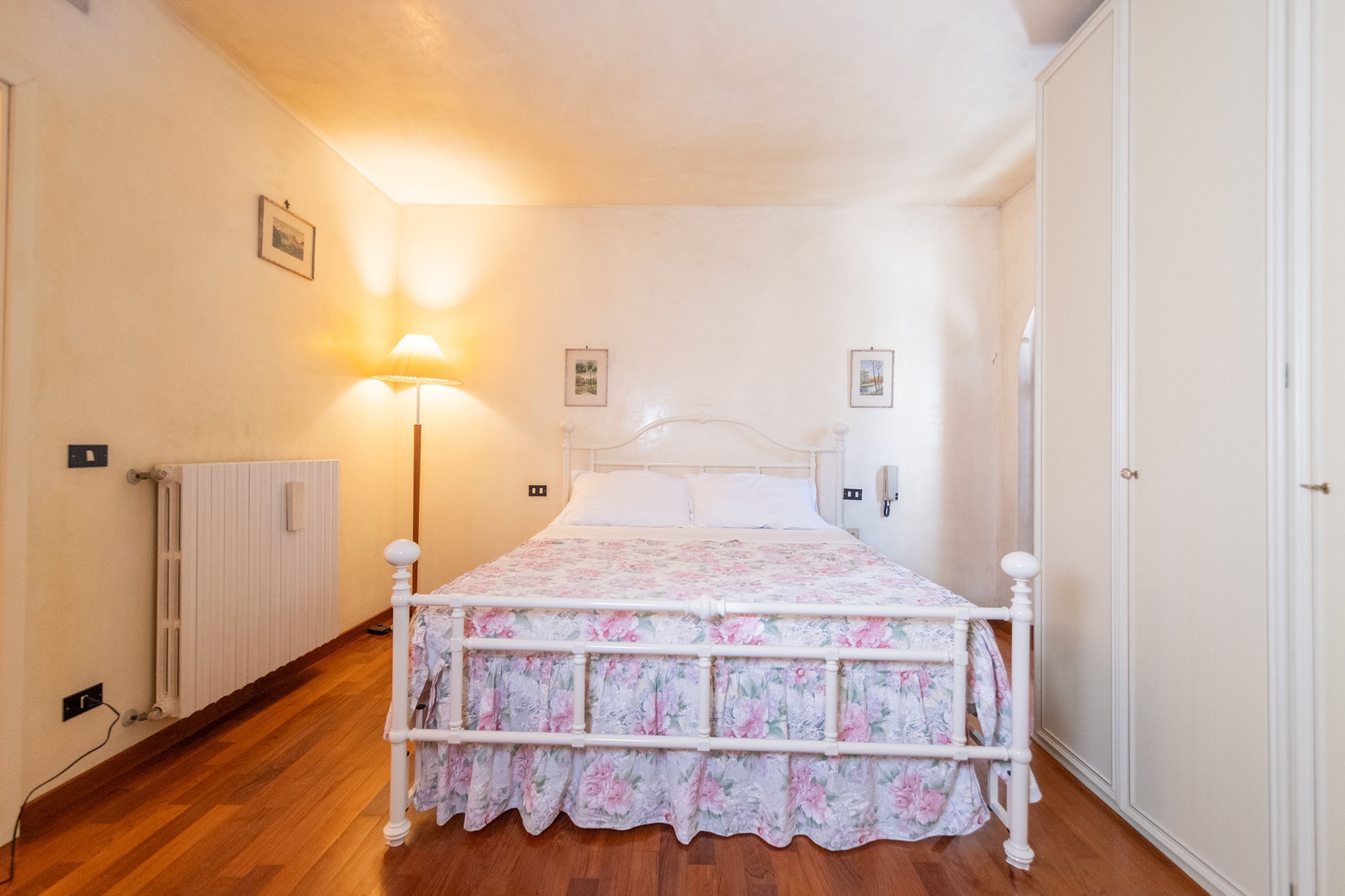 Une chambre avec un plancher en bois, un lit blanc recouvert d’une couvre-lit à motifs floraux, une lampe de chevet et un radiateur.