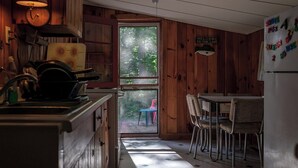 A cabin interior with wooden walls, a kitchen area with a sink and stove, a dining table with chairs, and a view of the outdoors through a door. - Cozy 2-bdrm, 3-season Adirondack cabin at tranquil Great Sacandaga Lake w/WiFi (Hadley)