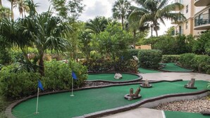 A miniature golf course with a green putting surface, surrounded by lush tropical vegetation and palm trees.