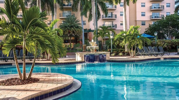 A swimming pool with a fountain, surrounded by palm trees and lounge chairs.