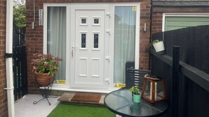 Una casa con puerta blanca, un pequeño jardín con una maceta y una mesa de cristal con otra maceta encima.