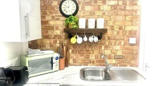 Una cocina con pared de ladrillo, un reloj, microondas, tostadora, lavabo y utensilios colgando.