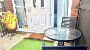 Un patio con una mesa de vidrio y dos sillas, una planta en maceta y una puerta con un cartel de advertencia.