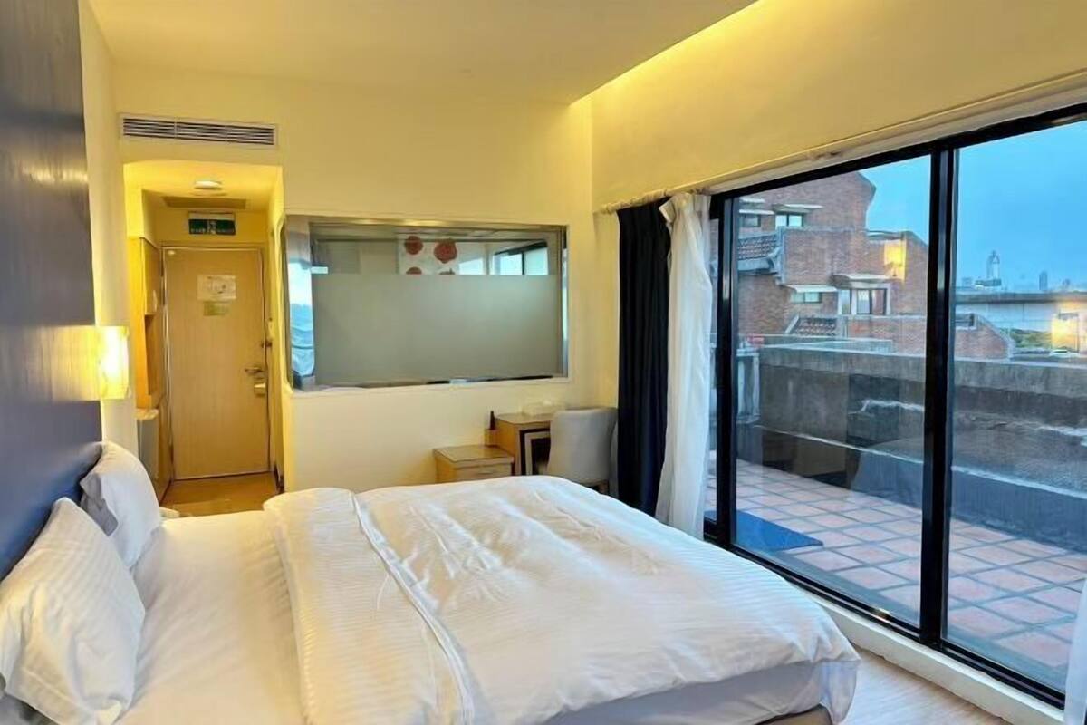 Une chambre d’hôtel avec un lit, un petit bureau, une fenêtre avec des rideaux et une vue sur les bâtiments environnants.