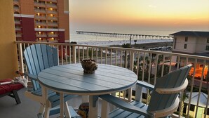Un balcón con una mesa redonda y sillas, con vista a un muelle y al océano al atardecer.