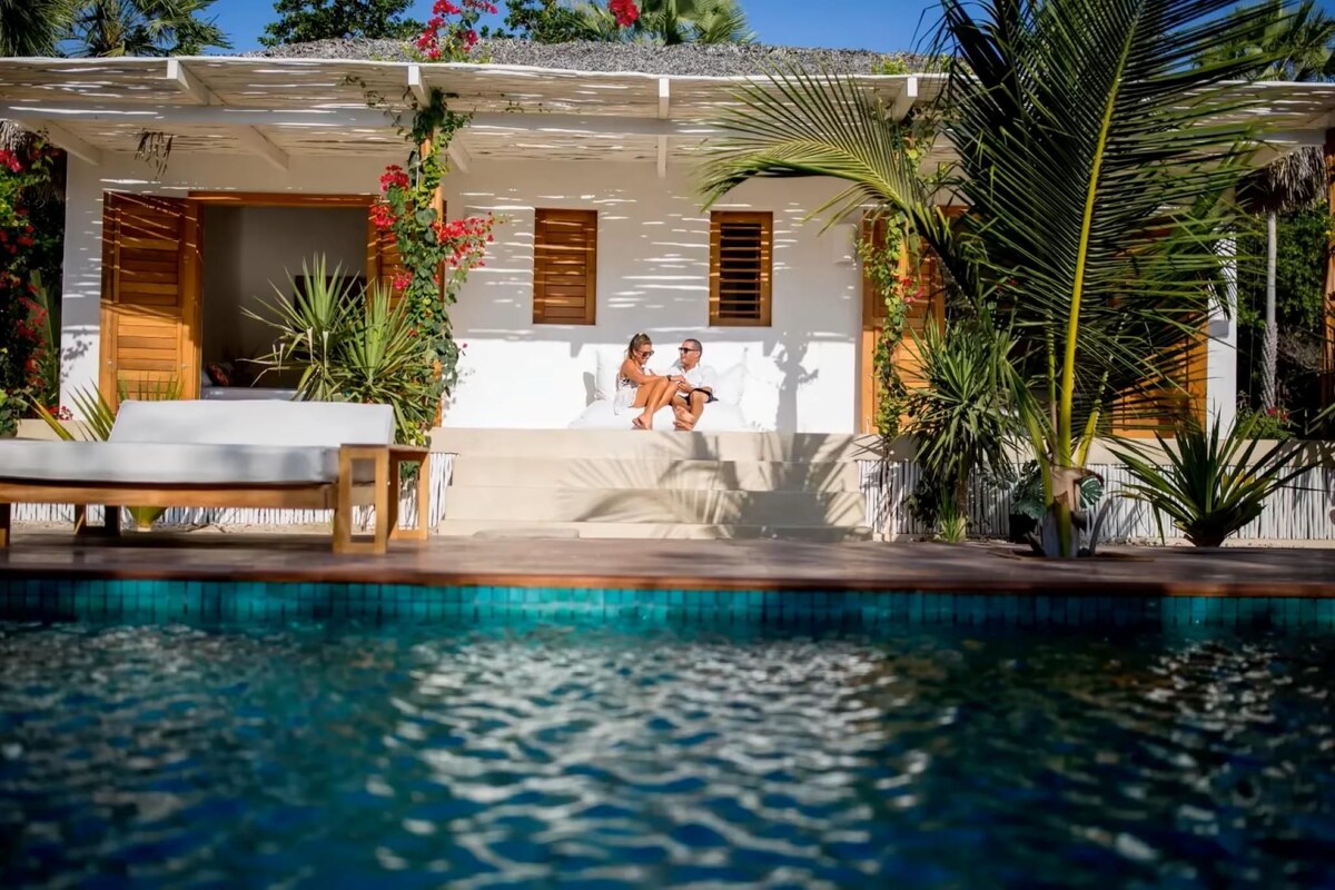 Zona junto a la piscina con una pareja sentada en un sofá blanco, rodeada de exuberante follaje y un edificio blanco con contraventanas de madera.