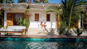 Un área junto a la piscina con una pareja sentada en un banco blanco, un edificio blanco con contraventanas de madera y plantas tropicales.