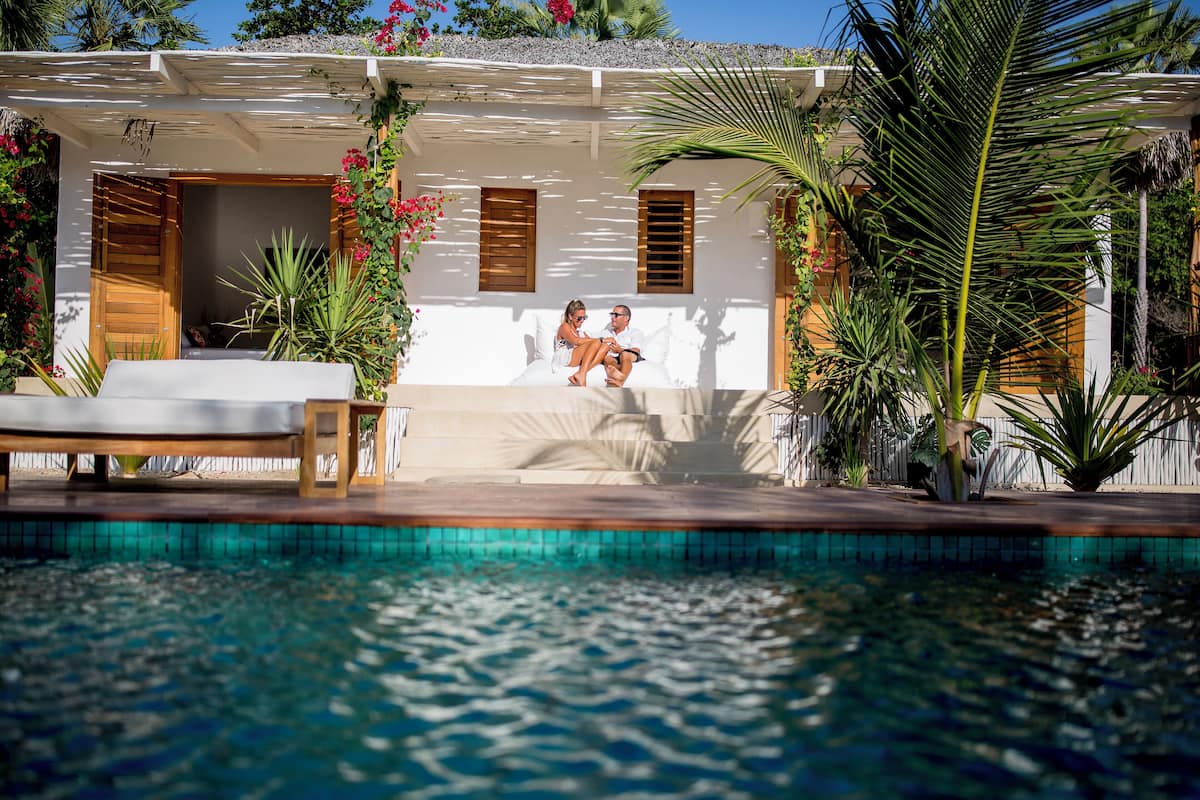 Un espace aménagé au bord de la piscine, avec un couple assis sur un banc blanc, un bâtiment blanc doté de volets en bois et des plantes tropicales.