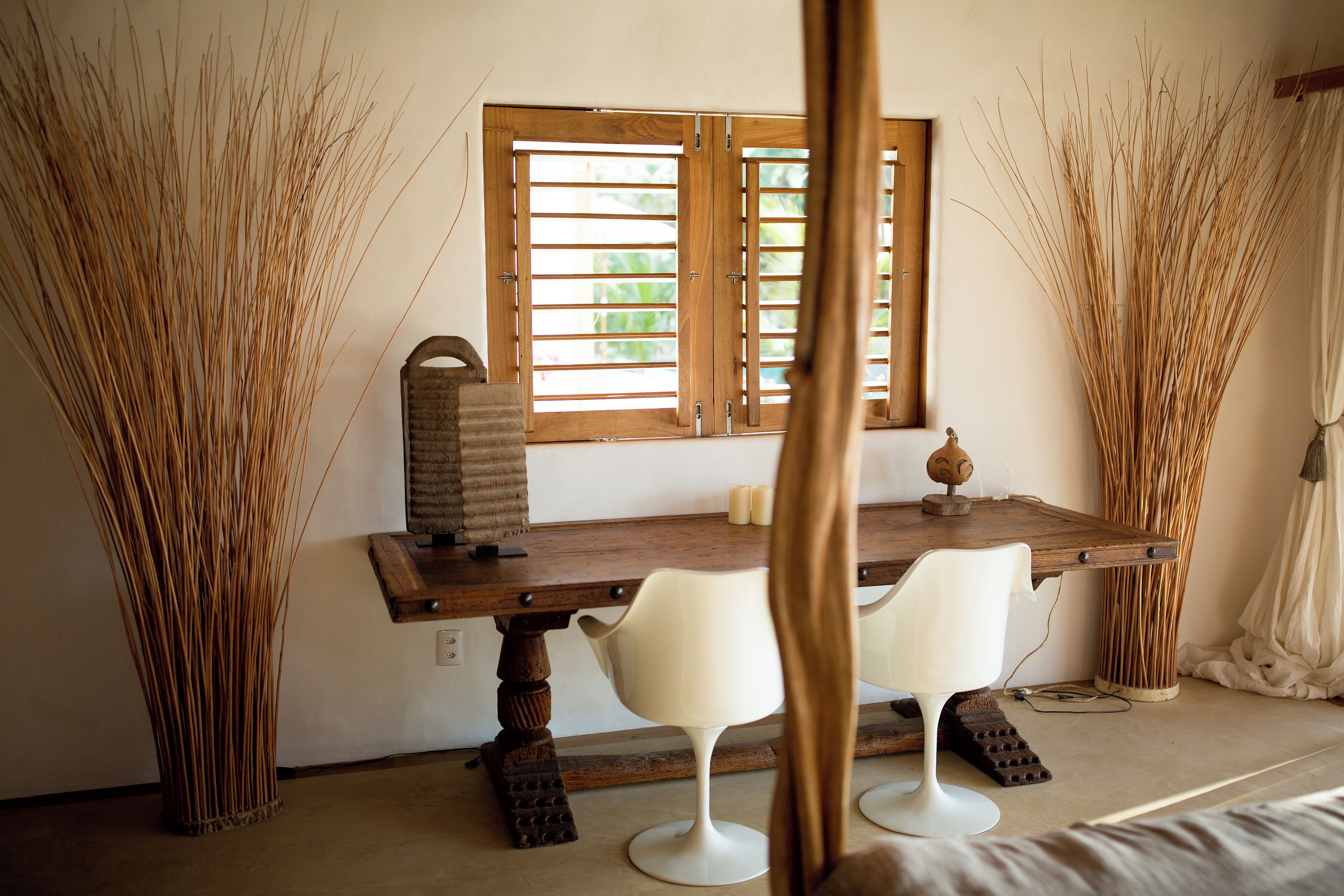 Una habitación con una mesa de madera, dos sillas blancas, una ventana con contraventanas de madera y altas plantas decorativas.