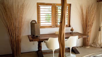 Una habitación con una mesa de madera, dos sillas blancas, una ventana con contraventanas de madera y altas plantas decorativas.