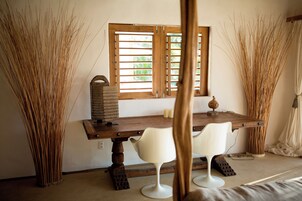 Una habitación con una mesa de madera, dos sillas blancas, una ventana con contraventanas de madera y altas plantas decorativas.