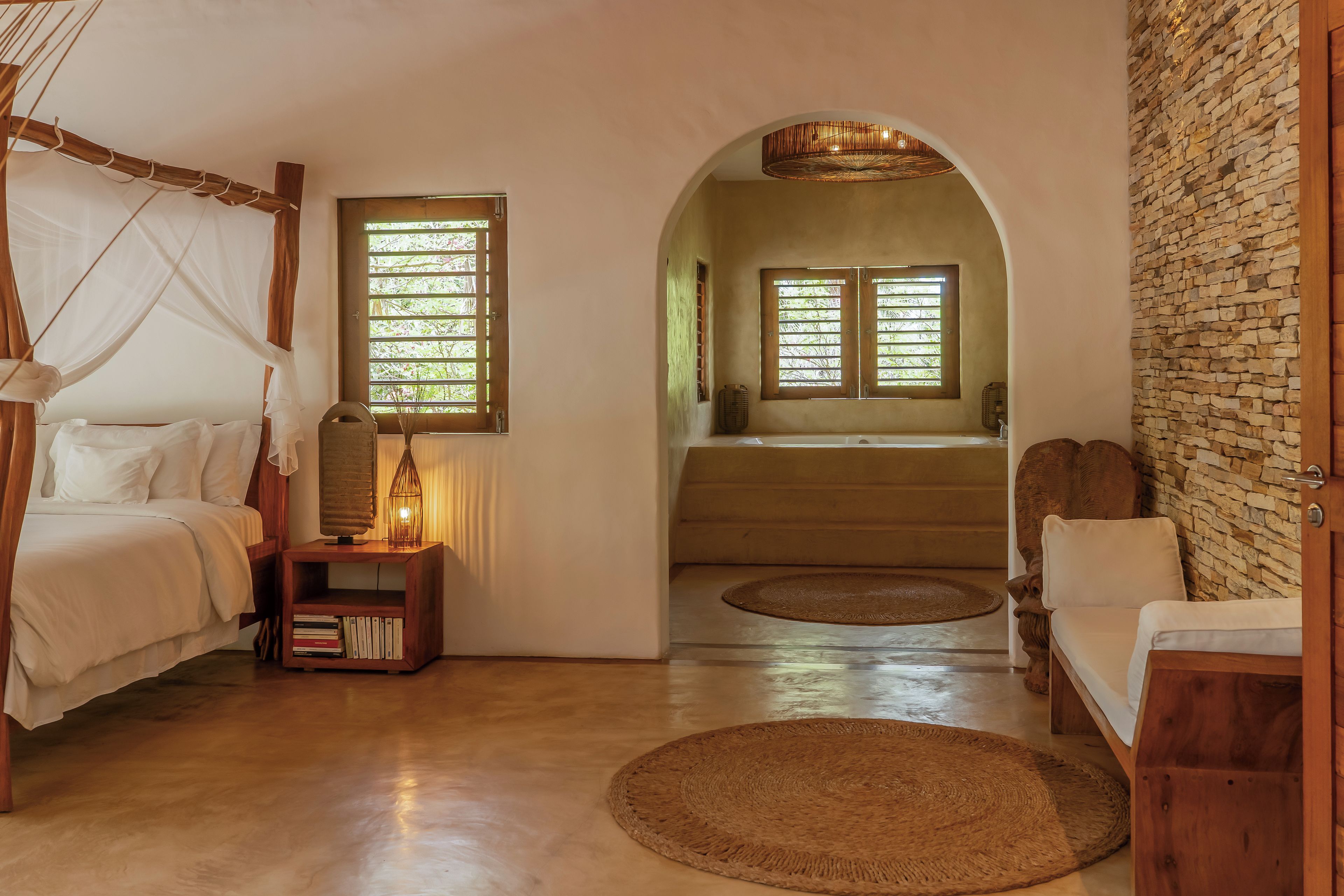 Un dormitorio con cama, mesita de noche, lámpara y vista a un baño a través de un arco.