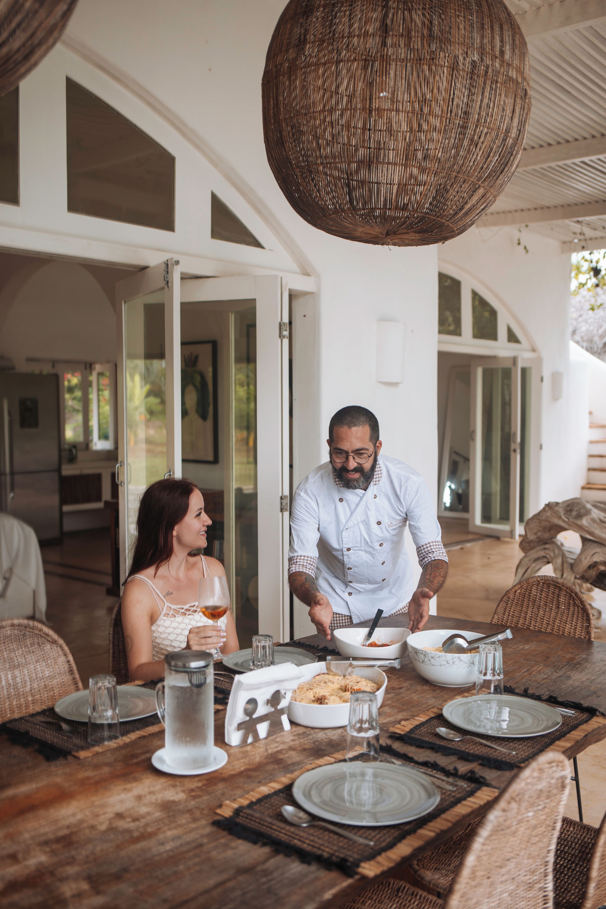 Un comedor con una mesa de madera dispuesta para dos personas, una lámpara colgante de mimbre y vista al interior a través de puertas de cristal.