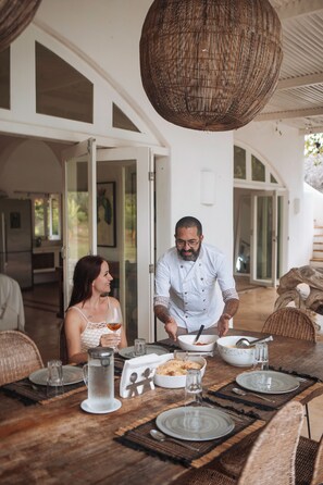 A dining area with a wooden table set for two, a wicker hanging light fixture, and a view of the interior through glass doors. - Villa Paz - La Villa Group (Camocim)