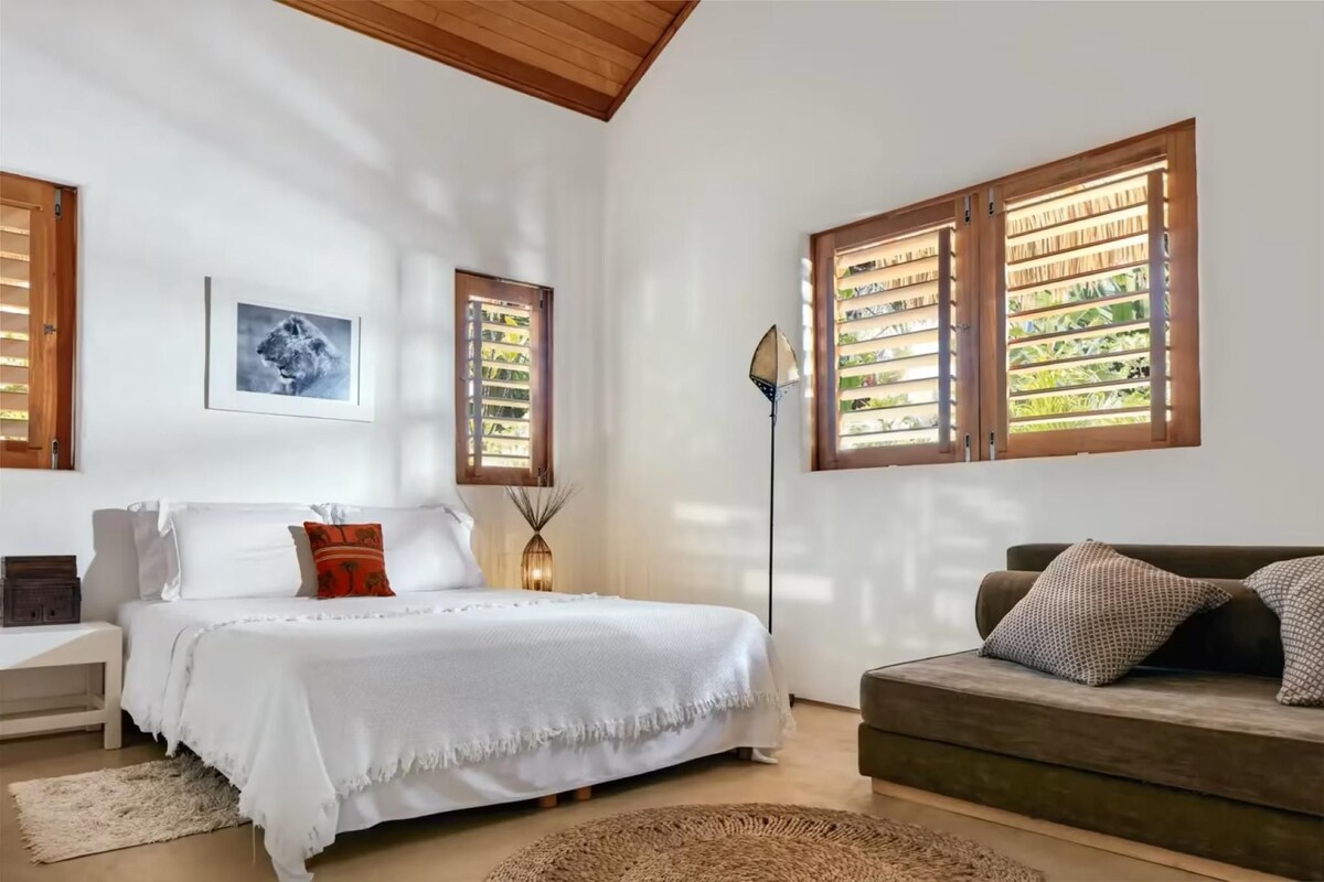A bedroom with a bed, a bedside table, a floor lamp, a sofa, and a window with blinds.