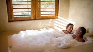 Una bañera llena de burbujas, una ventana con contraventanas de madera y dos copas de vino al borde.