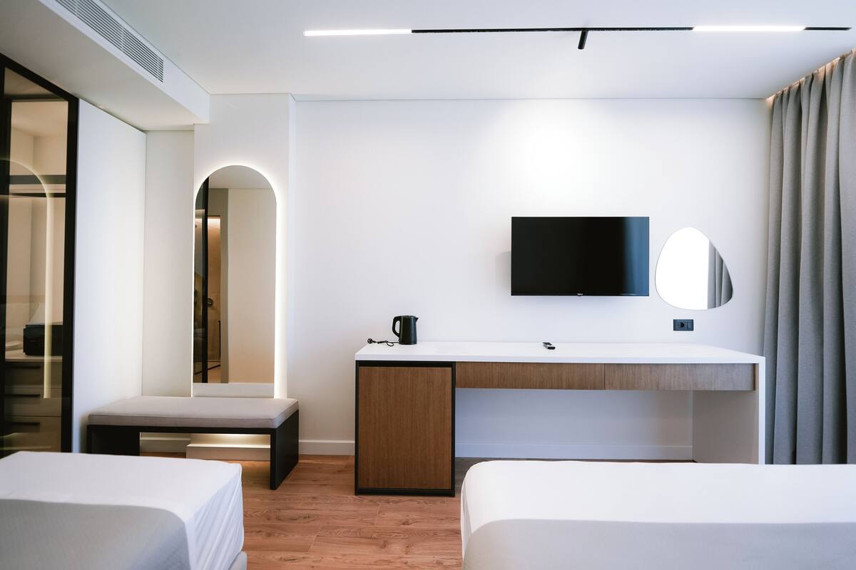 Une chambre d’hôtel moderne avec un lit blanc, un bureau en bois doté d’un téléviseur à écran plat, une table de chevet et un grand miroir.