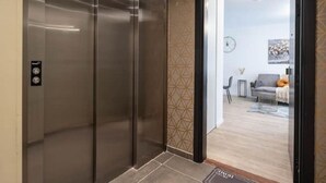 Un ascensor moderno con pantalla digital que conduce a una sala de estar con sofá, una mesa de centro y un elemento decorativo en la pared.