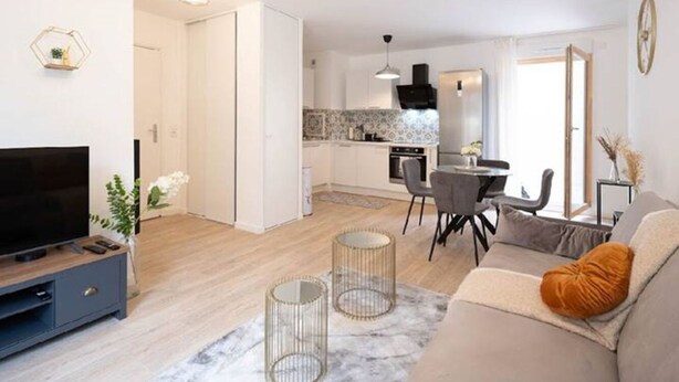 Una sala de estar moderna con sofá, televisor y objetos decorativos. Se ve una cocina al fondo.