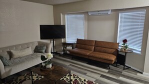 A living room with a beige sofa, a brown sofa, a coffee table, and a TV. There are two windows with blinds, an air conditioning unit, and a small table with a plant. - Marine View Townhouse (Everett)