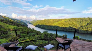 A wooden deck with two chairs and a table, overlooking a river and lush green hills. - COCOZNA Glamping (Samaná)