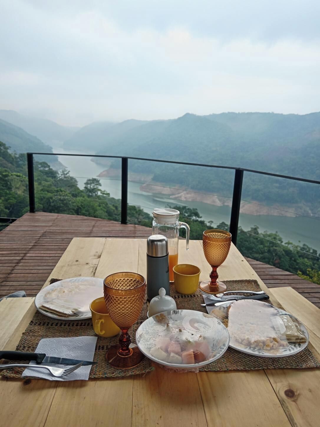 Ein Frühstückstisch auf einer Holzterrasse mit Blick auf einen Fluss und Berge.