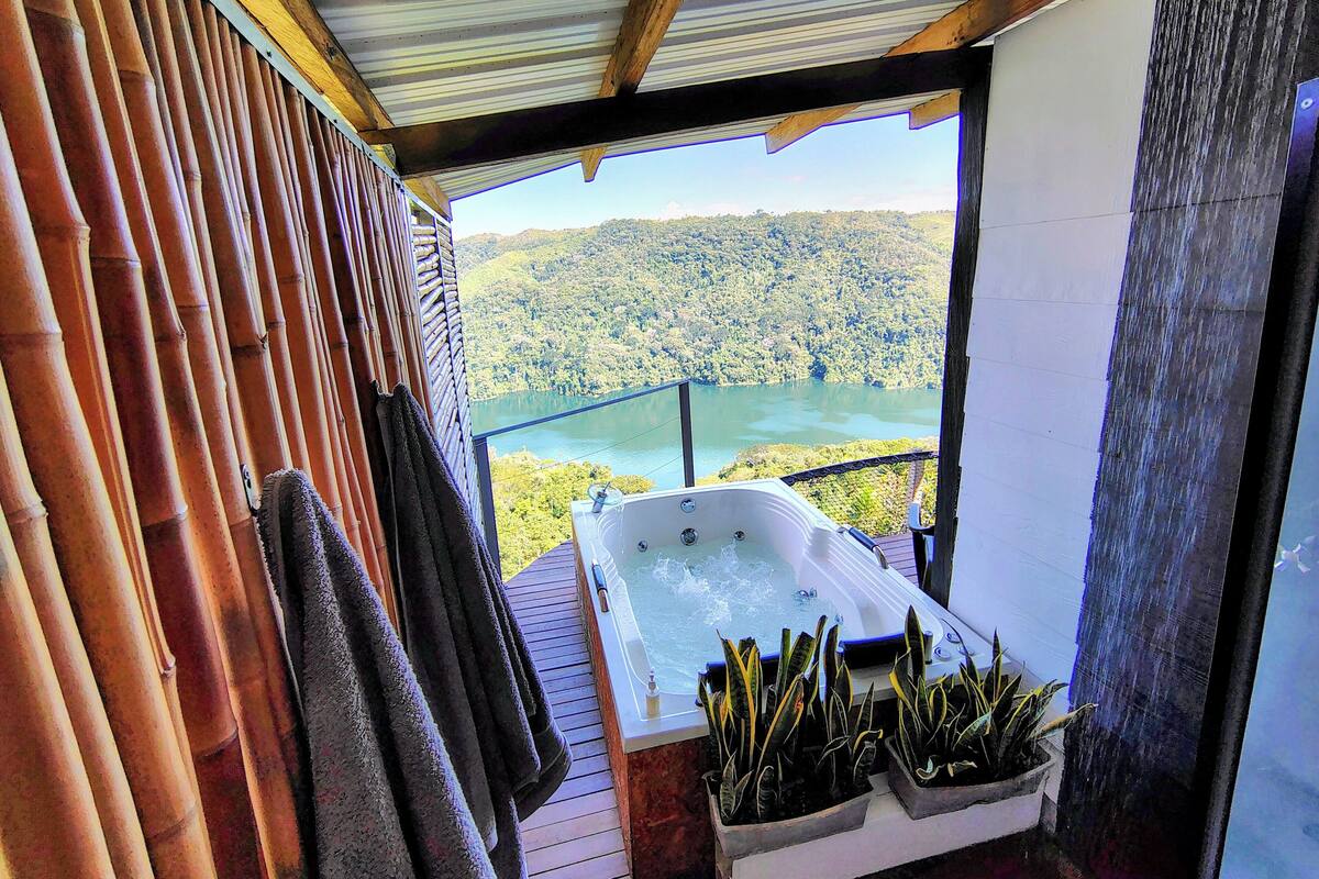 A balcony with a hot tub, bamboo wall, and a view of a lake and forest.