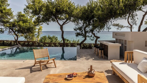 A poolside area with a view of the sea, a wooden table with fruits and a vase, and a sofa with cushions.