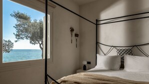 A bedroom with a bed, a bedside table, a nightstand, a window with a view of the sea, and a wall-mounted lamp.