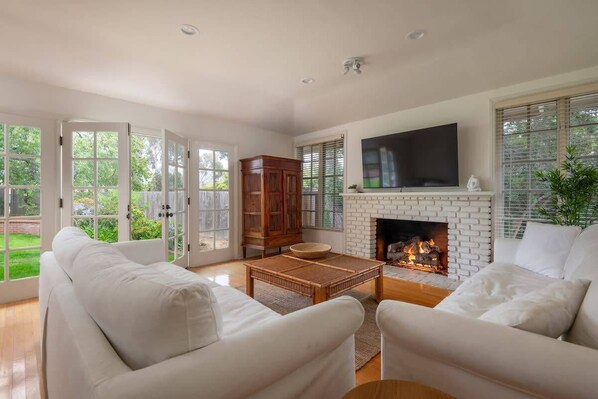 A spacious living room with a fireplace, white sofas, a flat-screen TV, and a large window overlooking a garden. - Casa Dorado (San Diego)