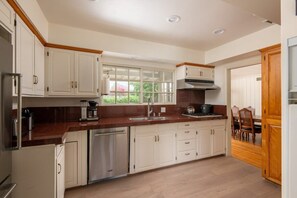 A kitchen with white cabinets, a stainless steel dishwasher, a coffee maker, and a window with a view of greenery.