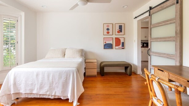 Une chambre à coucher comprenant un lit, des tables de chevet, un banc, un bureau avec une chaise, un ventilateur de plafond et des œuvres d’art accrochées au mur.