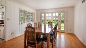 Une salle à manger avec une table et des chaises en bois, des murs recouverts de panneaux blancs et une vue sur un jardin, visible à travers des portes vitrées.