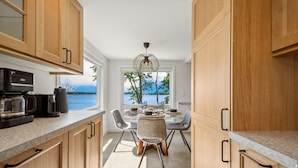 A modern kitchen with wooden cabinets, a coffee machine, and a dining area with a view of the water.