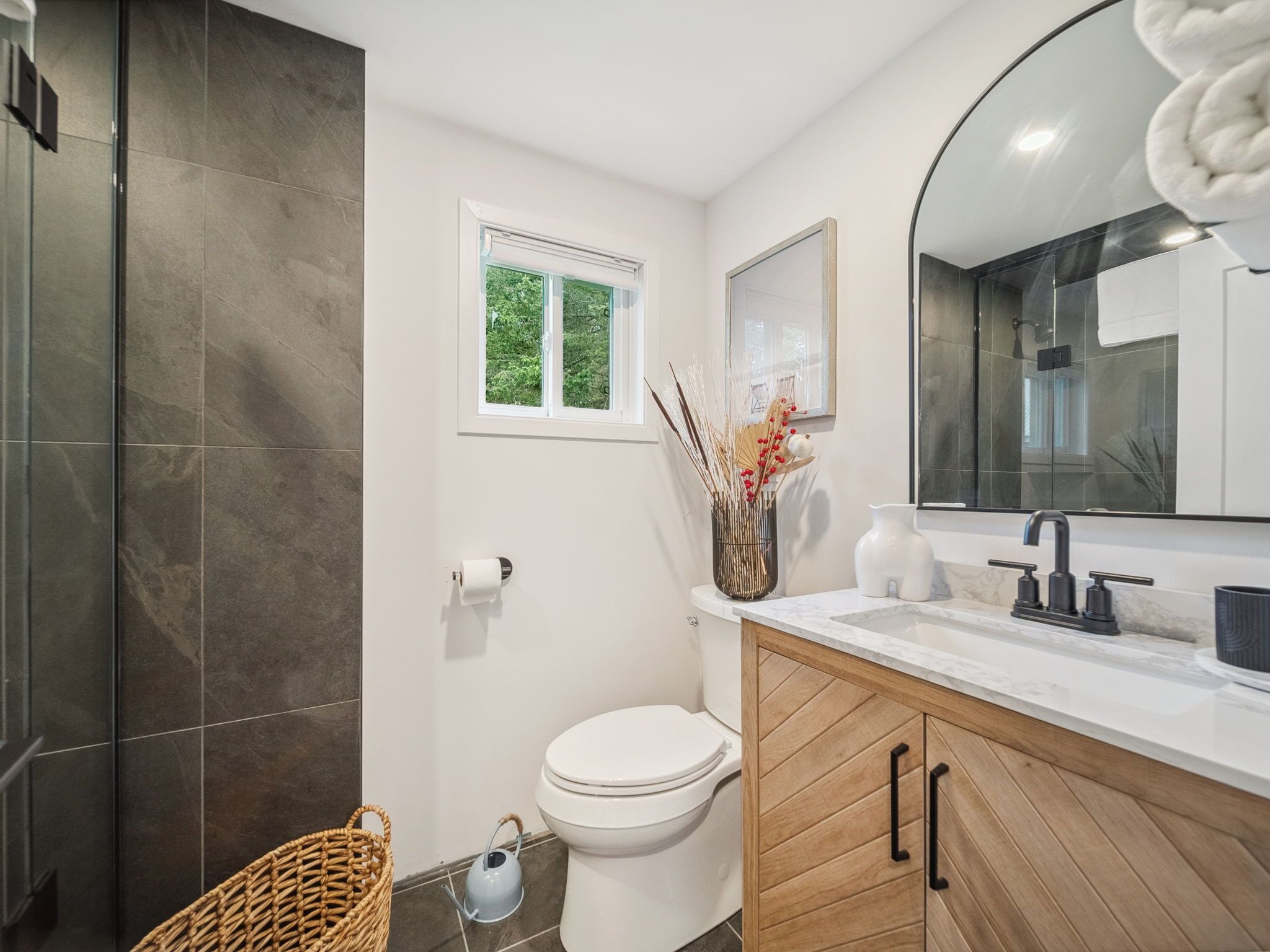 A modern bathroom with a toilet, sink, and shower. There is a window above the toilet, a mirror above the sink, and a towel hanging on the wall.
