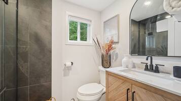 A modern bathroom with a toilet, sink, and shower. There is a window above the toilet, a mirror above the sink, and a towel hanging on the wall.