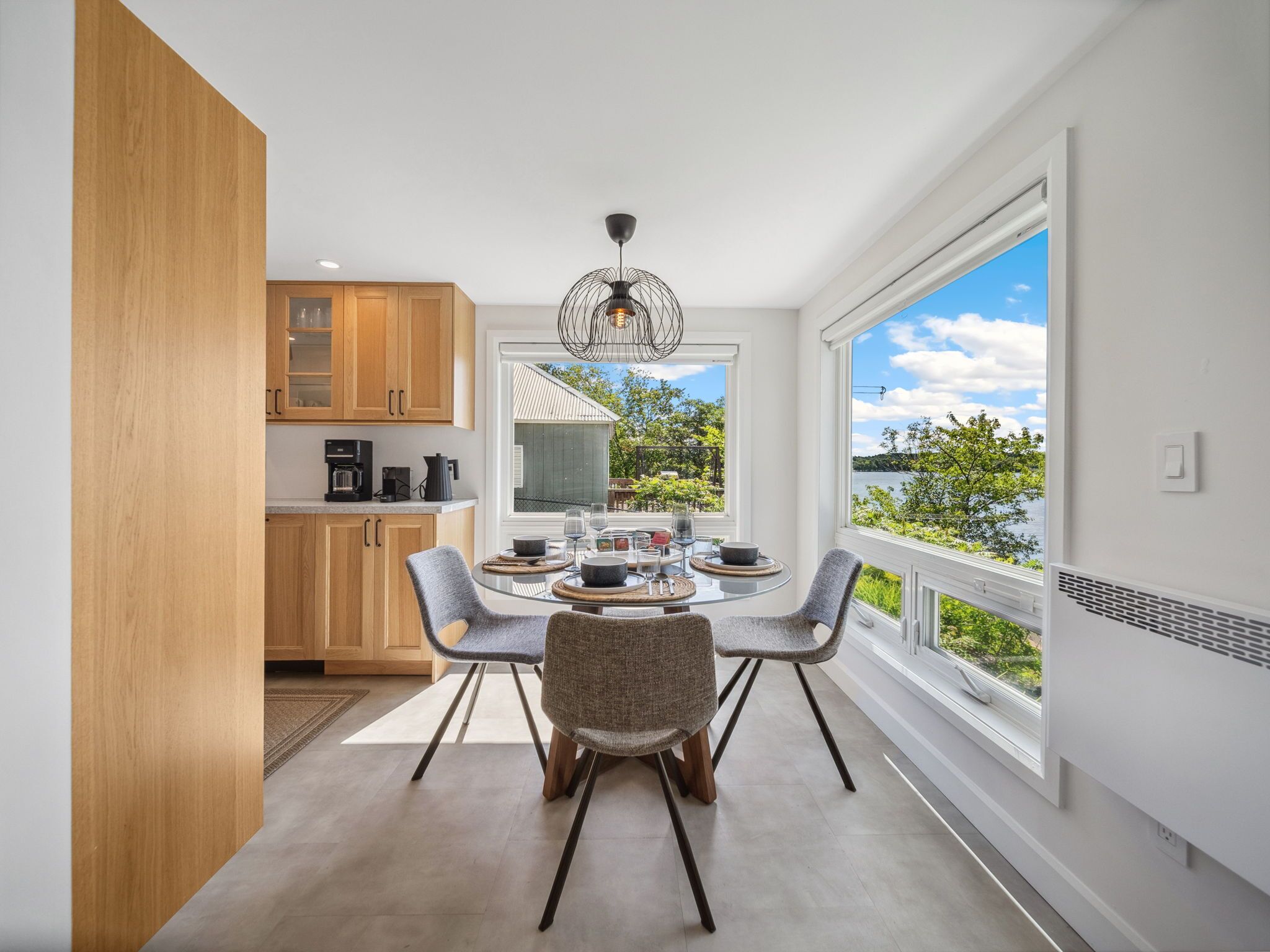 A modern kitchen with wooden cabinets, a dining area with a glass table and chairs, and a large window offering a view of a lake and greenery.