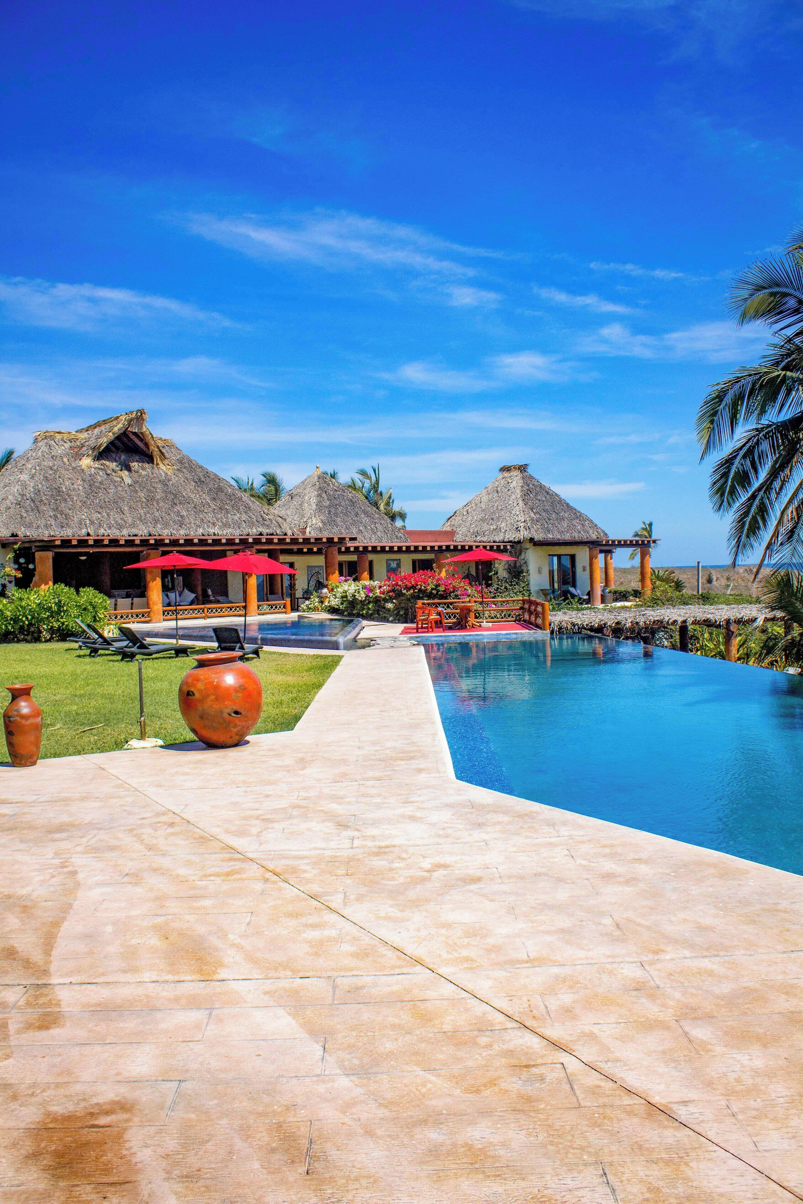 A resort with a swimming pool, thatched-roof structures, and a clear blue sky.