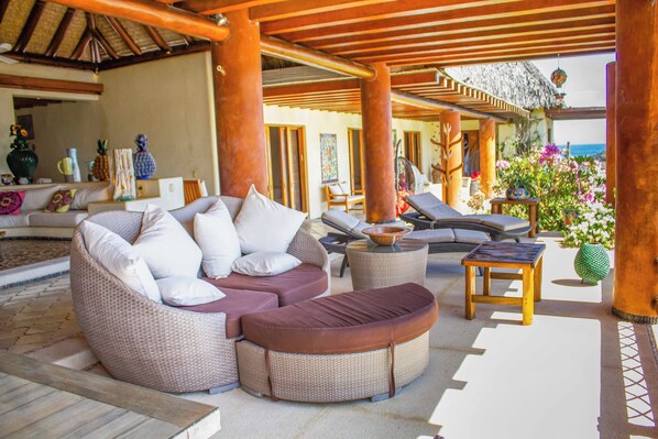 A covered outdoor seating area with wicker furniture, a wooden table, and a view of the ocean.