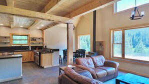 Amplia sala de estar con sofá de cuero, cocina con armarios de madera y grandes ventanales con vistas a un bosque.