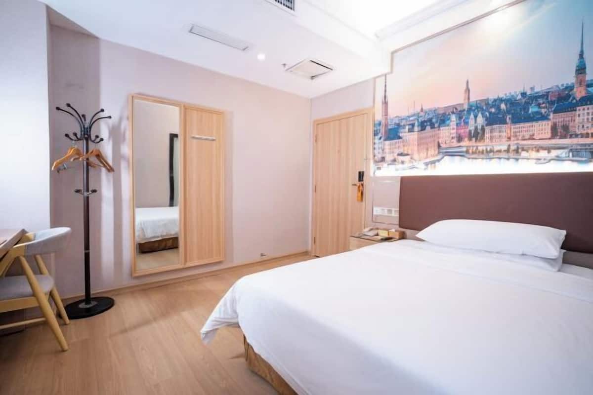 A hotel room with a bed, a desk, a coat rack, and a large window with a cityscape view.
